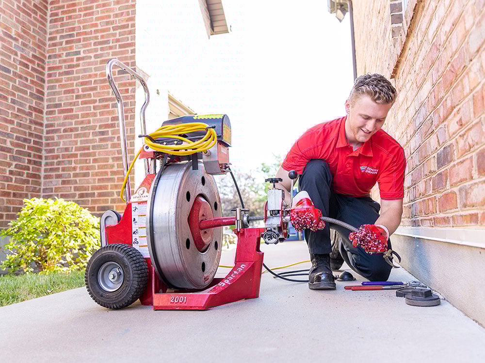 A technician wearing a red uniform kneels outside a brick building, operating a drain-cleaning machine and hose on a concrete walkway to clear a pipe.