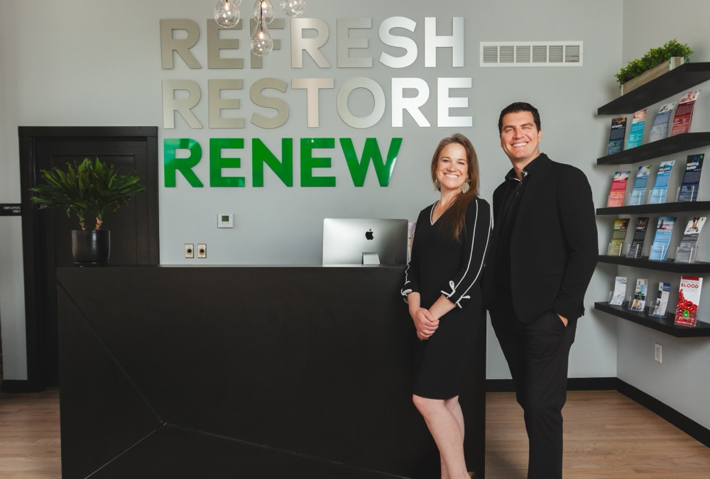 Two adults stand smiling behind a modern reception desk in a clean, contemporary clinic lobby. Large wall lettering reads “REFRESH RESTORE RENEW,” with “RENEW” in green. A computer sits on the desk, pendant lights hang overhead, and shelves with brochures line the wall to the right.