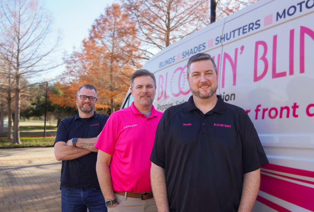 Three men stand outdoors in front of a branded service van with pink lettering for a blinds and shutters company. They wear black and pink branded polo shirts and smile at the camera, with autumn trees and a paved walkway in the background.
