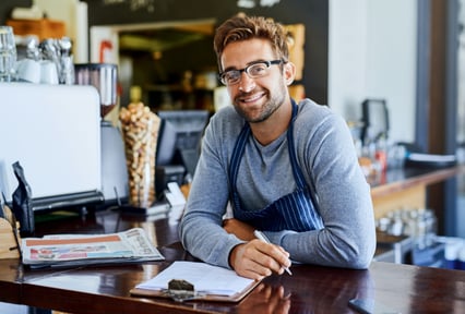 A smiling man in an apron with a clip board leaning on a counter