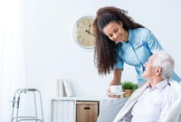 Home healthcare caregiver serving a cup of tea to an elderly man sitting on a couch.