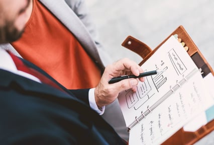 A man's hand holding a pen pointing at data in a planner