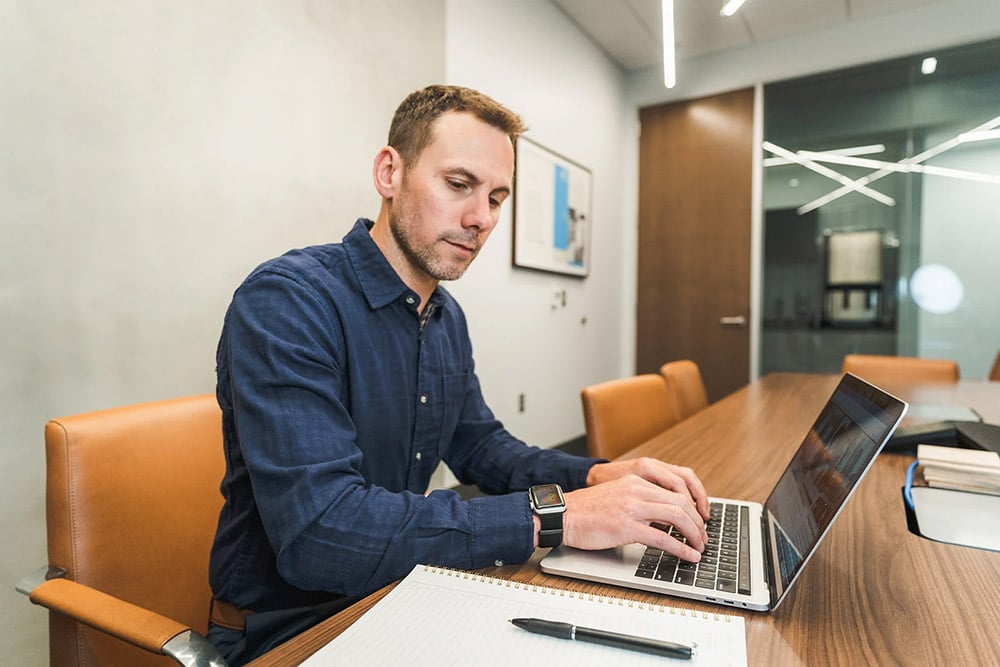 A man sits at a conference table working on a laptop, with a notebook and pen beside him. He wears a blue button-down shirt and focuses on typing in a modern office meeting room with glass walls and leather chairs.