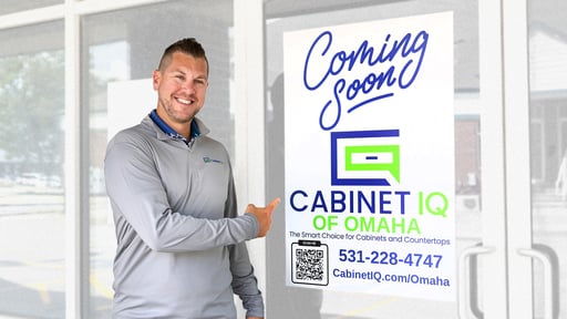 A smiling man wearing a light gray long-sleeve shirt points to a large “Coming Soon” sign on a glass door. The sign displays the Cabinet IQ logo and reads “CABINET IQ of Omaha – The Smart Choice for Cabinets and Countertops,” along with a QR code, phone number, and website URL.