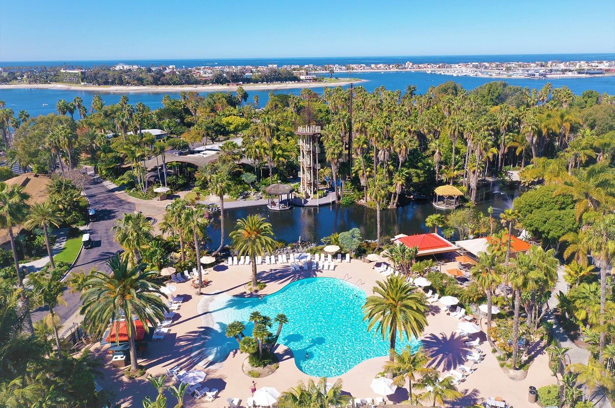 Aerial view of a tropical resort with a large outdoor pool surrounded by palm trees and lounge chairs, set beside a lagoon with the ocean and coastline visible in the distance.