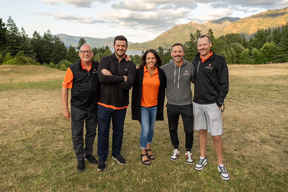 Six advisors standing side by side on a grassy field, smiling at the camera, wearing matching orange and black branded apparel, with trees, a lake, and mountains in the background.