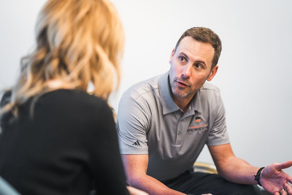 A business advisor in a gray polo shirt speaks with a client in a one-on-one meeting, gesturing as they discuss options in a professional office setting.