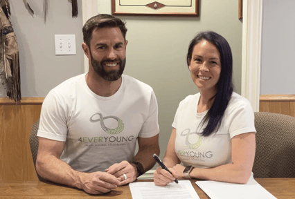 A man and a woman sit at a table smiling at the camera, both wearing matching white “4EverYoung Anti-Aging Solutions” T-shirts. The woman is signing a document with a pen while the man rests his hands on the table. They appear to be in an office setting with wood paneling and framed décor on the walls.