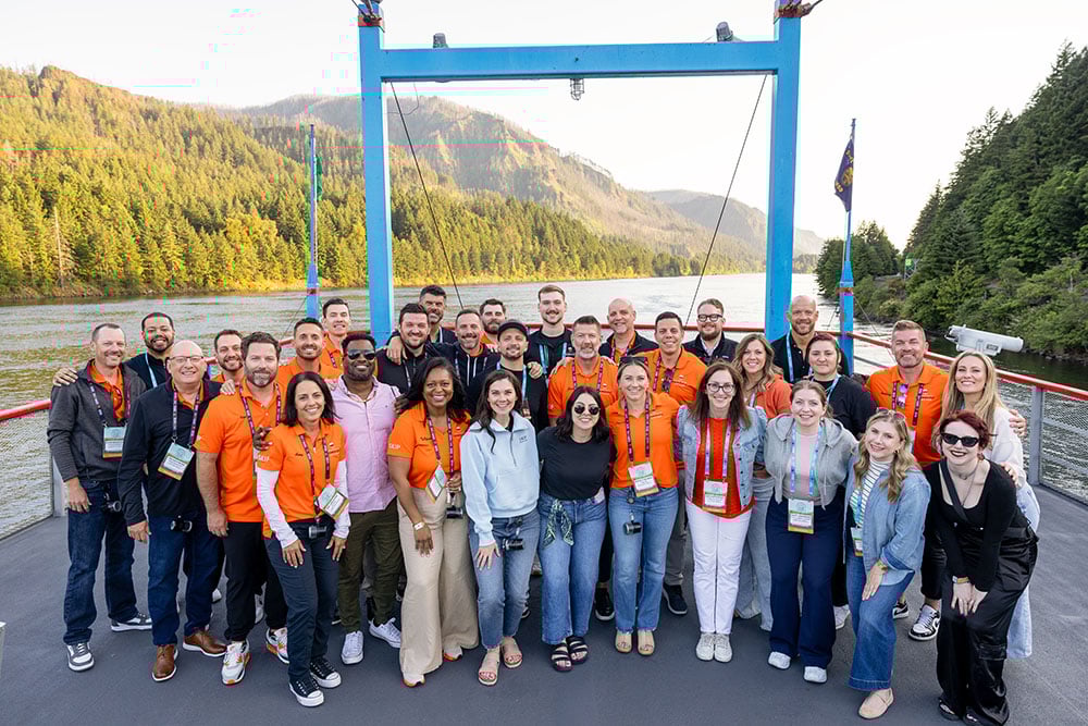 A large group of adults poses together on a boat deck, many wearing matching orange shirts and event badges. Forested hills and a river surround them, creating a scenic outdoor setting that suggests a team outing or company retreat.