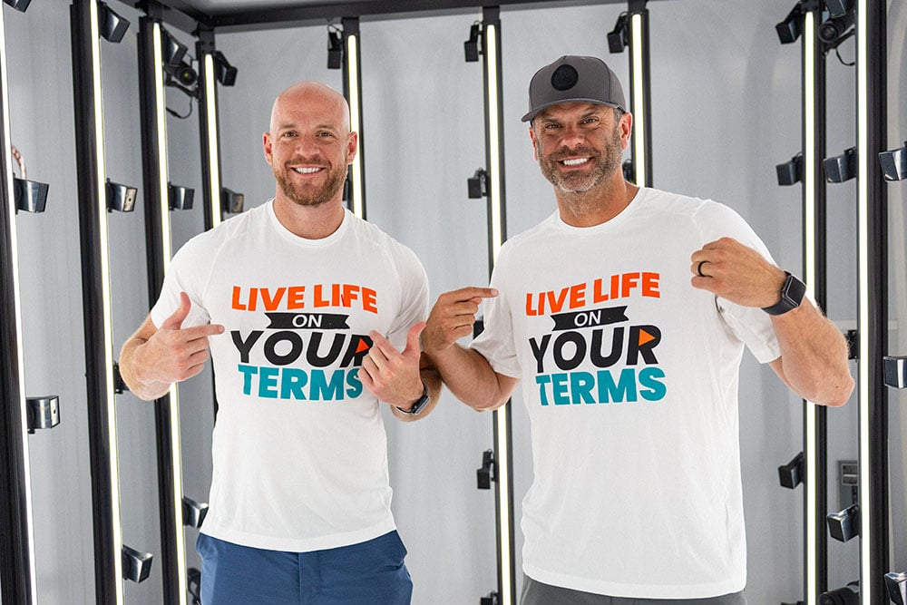 Two men stand smiling in a studio setting, pointing to matching white T-shirts that read “Live Life on Your Terms.”