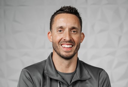 A smiling man with short dark hair and a trimmed beard wearing a gray zip-up jacket, posed against a light gray textured background.