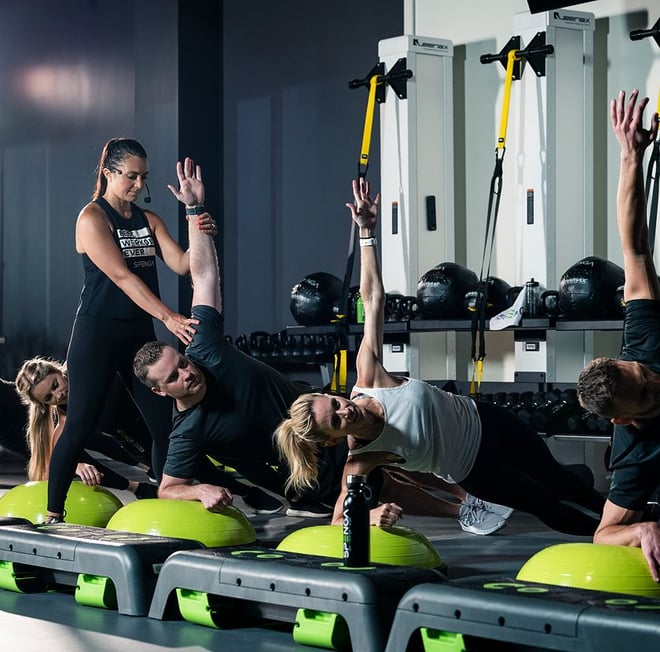Inside the SPENGA fitness studio, an instructor helps adjust a client's form, while a group of five client's are doing a strength exercise.