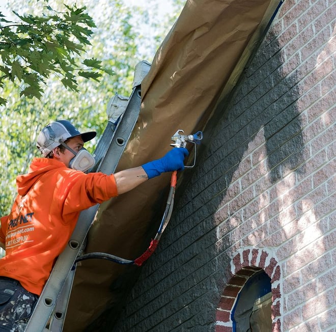 Spray-Net employee stands on ladder leaned against brick house, spraying over brick with dark grey paint.