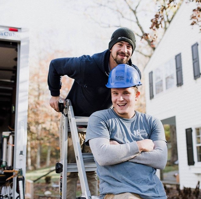 Two Brothers Gutters employees stand smiling on a ladder in front of a white and black house.