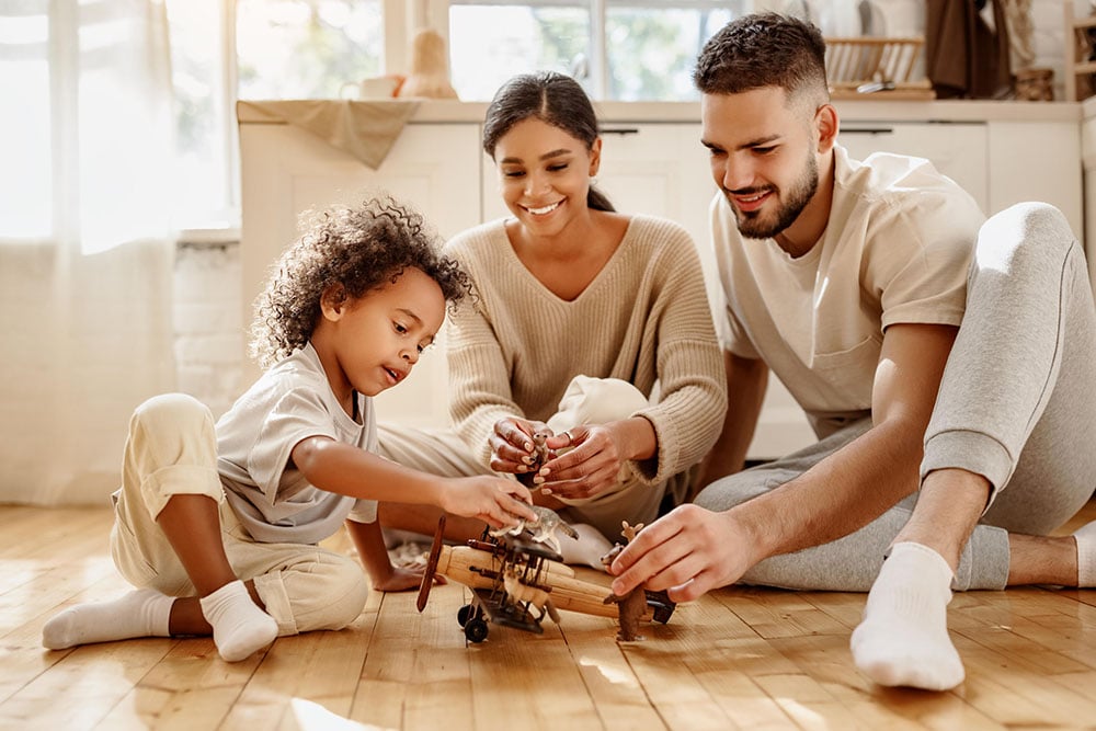 Smiling parents sit on a wooden floor with their young child, playing together with a toy airplane in a bright, cozy kitchen filled with natural light.