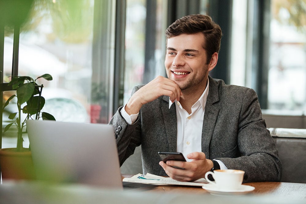 Smiling man in a blazer sits at a café table with a laptop and smartphone, holding his phone while looking off to the side, with a cup of coffee in front of him.