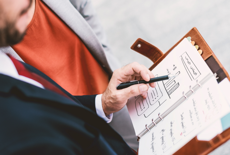 A man's hand holding a pen pointing at data in a planner