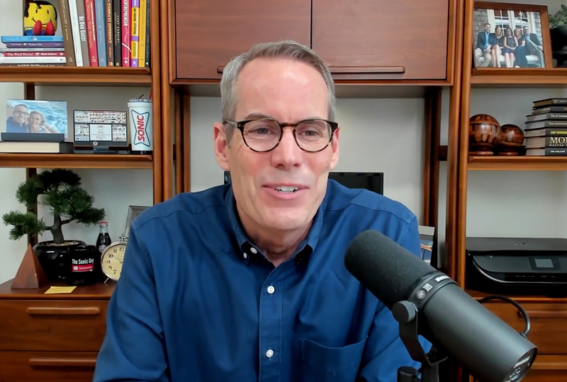 A man wearing glasses speaks into a microphone during a podcast recording, seated at a desk with bookshelves and office items behind him.