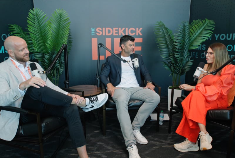 Three people seated in armchairs recording a podcast with microphones in a studio, with a backdrop that reads ‘The Sidekick Live’ and large plants on either side.
