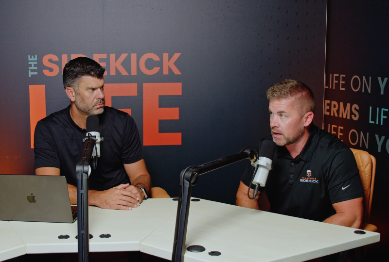 Two men speaking into microphones while recording a podcast at a table, with a ‘The Sidekick Live’ backdrop behind them.