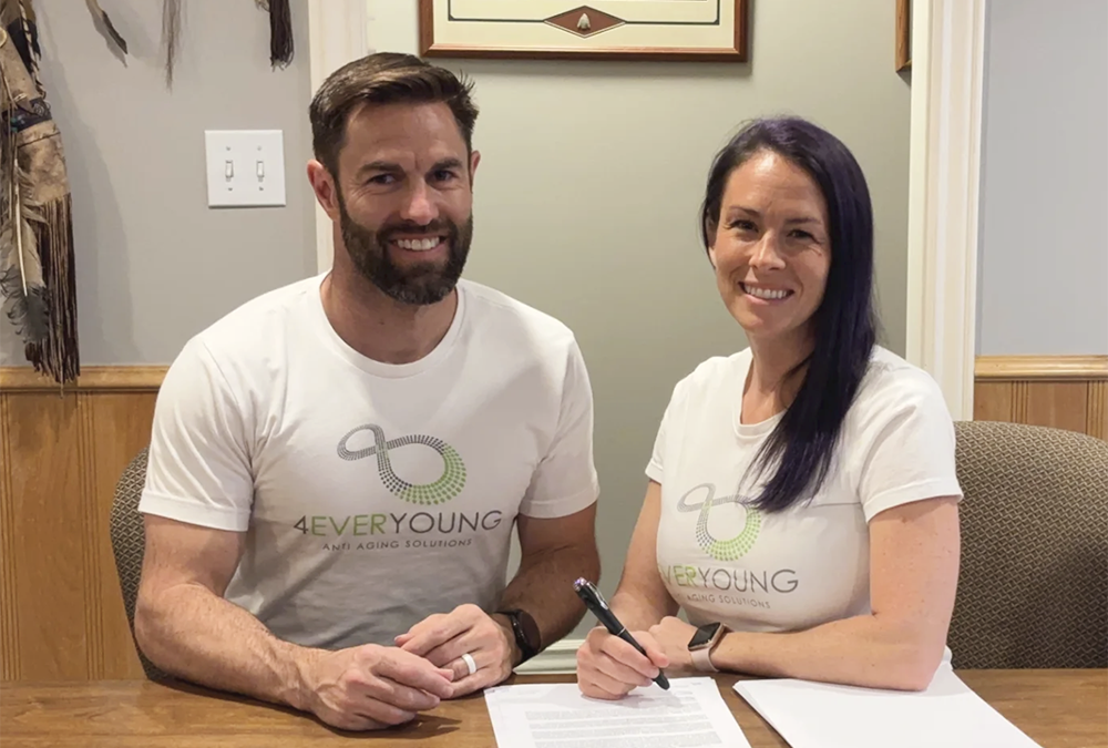 A man and a woman sit at a table smiling at the camera, both wearing matching white “4EverYoung Anti-Aging Solutions” T-shirts. The woman is signing a document with a pen while the man rests his hands on the table. They appear to be in an office setting with wood paneling and framed décor on the walls.