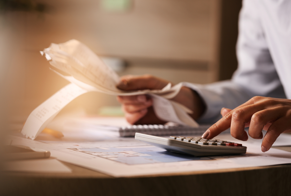 Close-up of a person reviewing long receipts and pressing buttons on a calculator at a desk covered with paperwork.