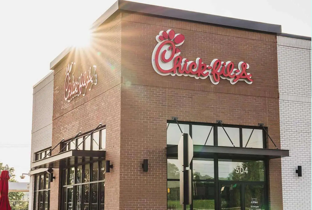 A Chick-fil-A restaurant building with brick exterior and large red Chick-fil-A logo on the front. Sunlight shines from behind the roof, creating a bright flare. The entrance features glass windows and black awnings.
