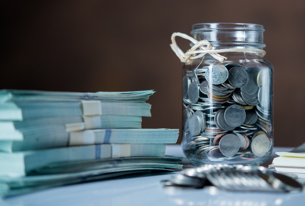 A glass jar filled with assorted coins sits on a reflective surface beside several neatly stacked bundles of paper money, with additional loose coins in the foreground.