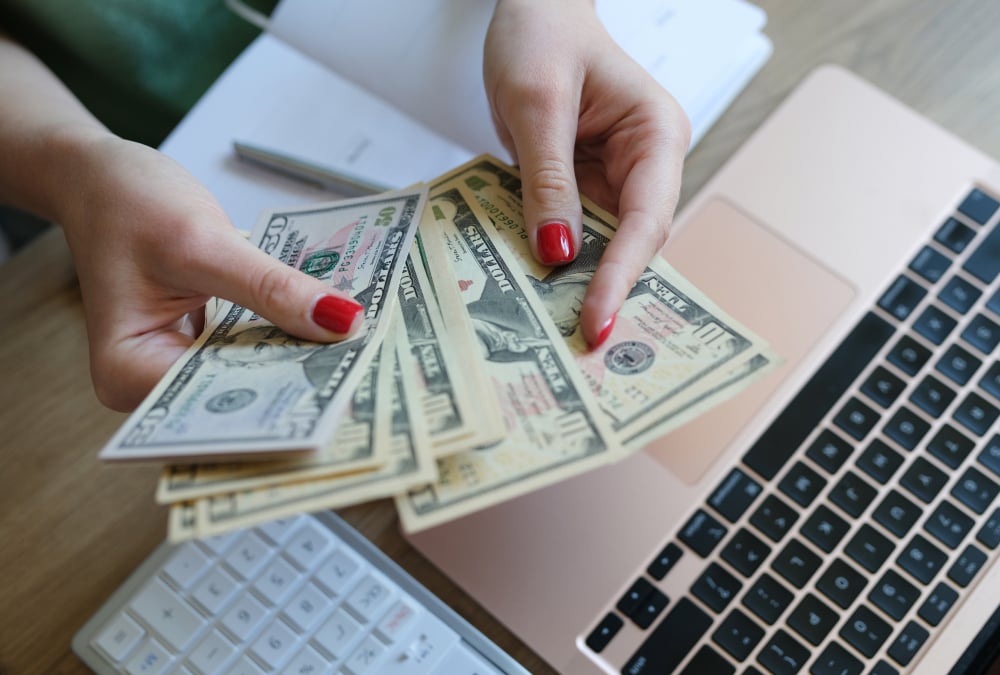 A person with red-painted fingernails holds several U.S. dollar bills in both hands over a laptop keyboard on a desk, with a calculator and papers nearby.