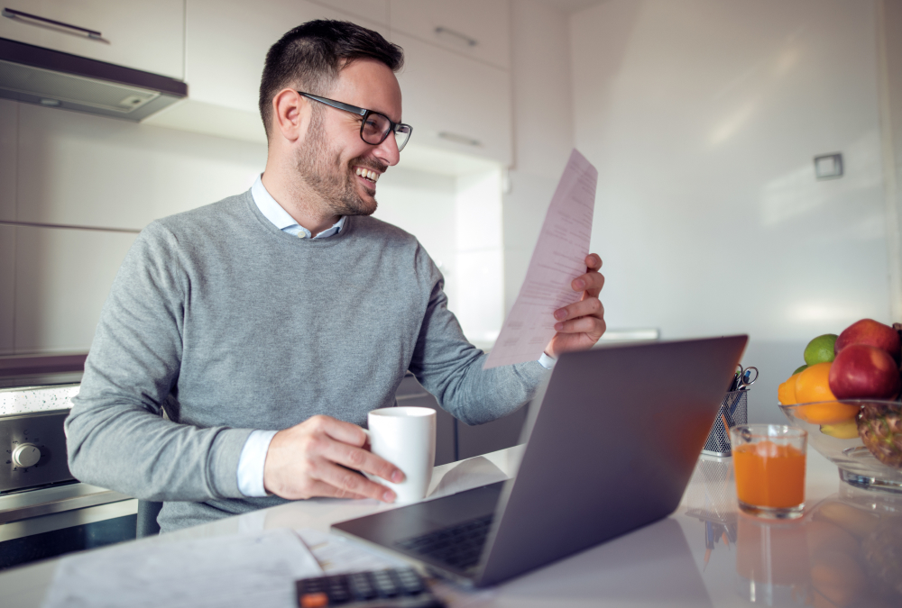 A guy sitting in front of a laptop holding a piece of paper and a mug, smiling